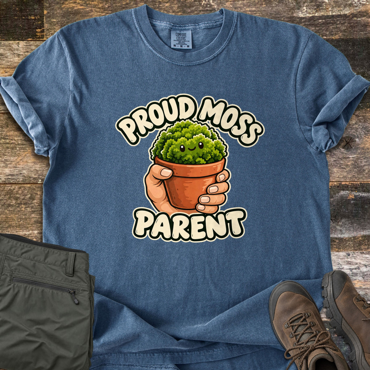 Proud Moss Parent Comfort Colors T-shirt in Denim, laid flat. A smiling cartoon moss clump in a terracotta pot, held in a hand, with the words Proud Moss Parent. It lies on weathered wood planks beside folded olive hiking pants and brown hiking boots.