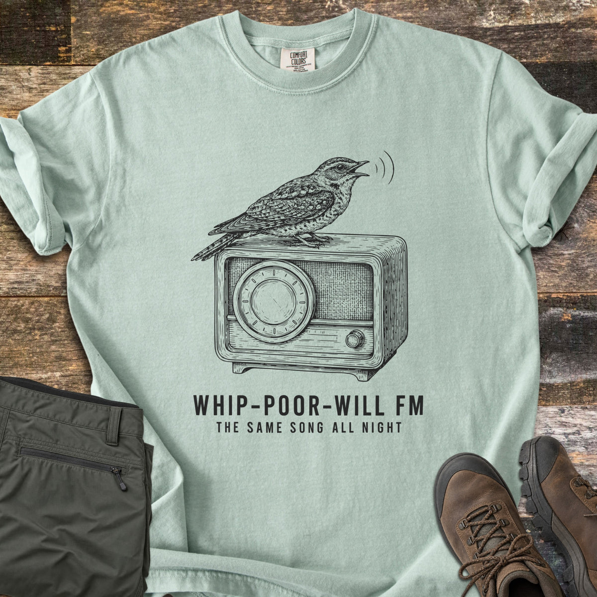 Whip-Poor-Will FM Comfort Colors T-shirt in Bay, laid flat. A sketched whip-poor-will sings atop a vintage radio above the words Whip-Poor-Will FM, The Same Song All Night. It lies with rolled sleeves on weathered wood beside folded olive pants and brown hiking boots.