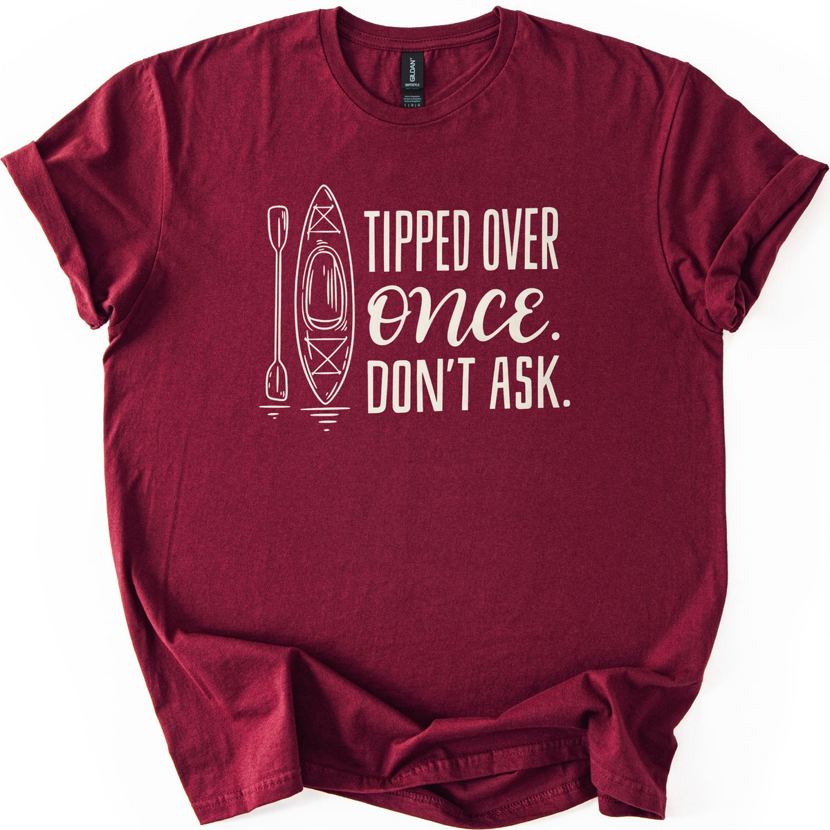 Tipped Over Once Kayak Comfort Colors T-shirt in Maroon, laid flat. A cream line drawing of an upright kayak and paddle with water ripples sits beside the words Tipped Over Once. Don't Ask., with Once in script. The shirt has cuffed sleeves on a white background.