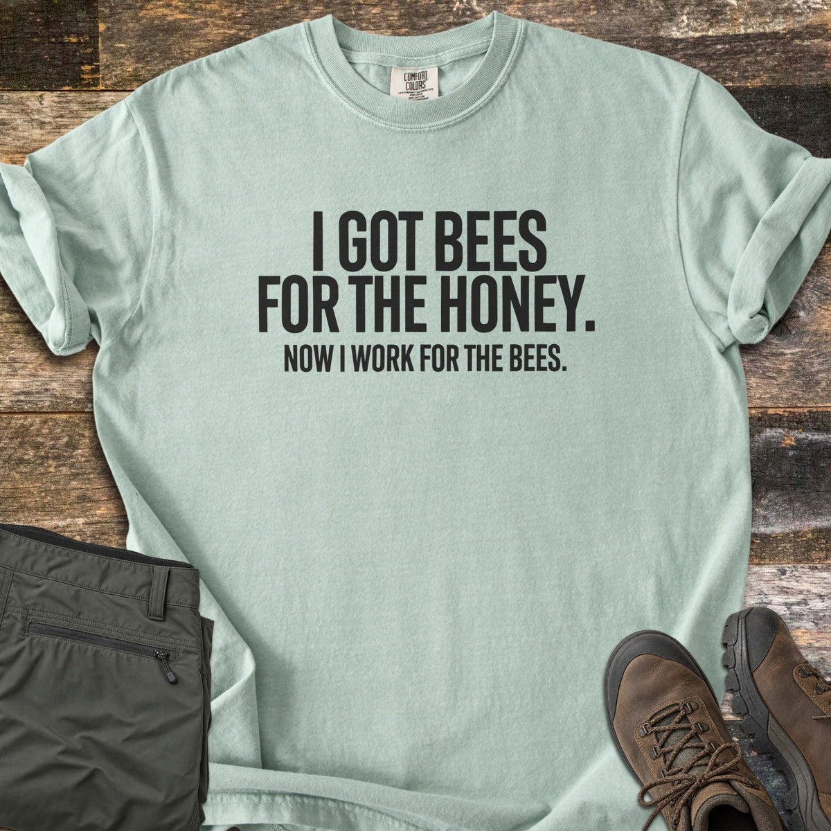 Work For The Bees Comfort Colors T-shirt in Bay, laid flat. Black block letters read I GOT BEES FOR THE HONEY. NOW I WORK FOR THE BEES. The shirt lies with cuffed sleeves on weathered wood planks beside folded olive green pants and brown hiking boots.