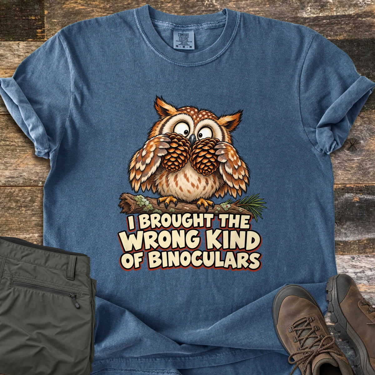 I Brought The Wrong Kind Of Binoculars Comfort Colors T-shirt in Denim, laid flat. A wide-eyed cartoon owl on a mossy branch holds two pinecones to its eyes like binoculars above the words I Brought The Wrong Kind Of Binoculars. Olive hiking pants and brown boots sit on weathered wood.