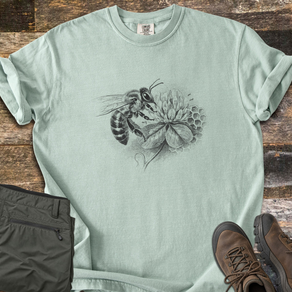 Honeybee On Clover Comfort Colors T-shirt in Bay, laid flat. A detailed black engraving-style illustration of a honeybee on a clover blossom with honeycomb cells behind it, no words. Sleeves rolled, on weathered wood planks beside olive hiking pants and brown hiking boots.