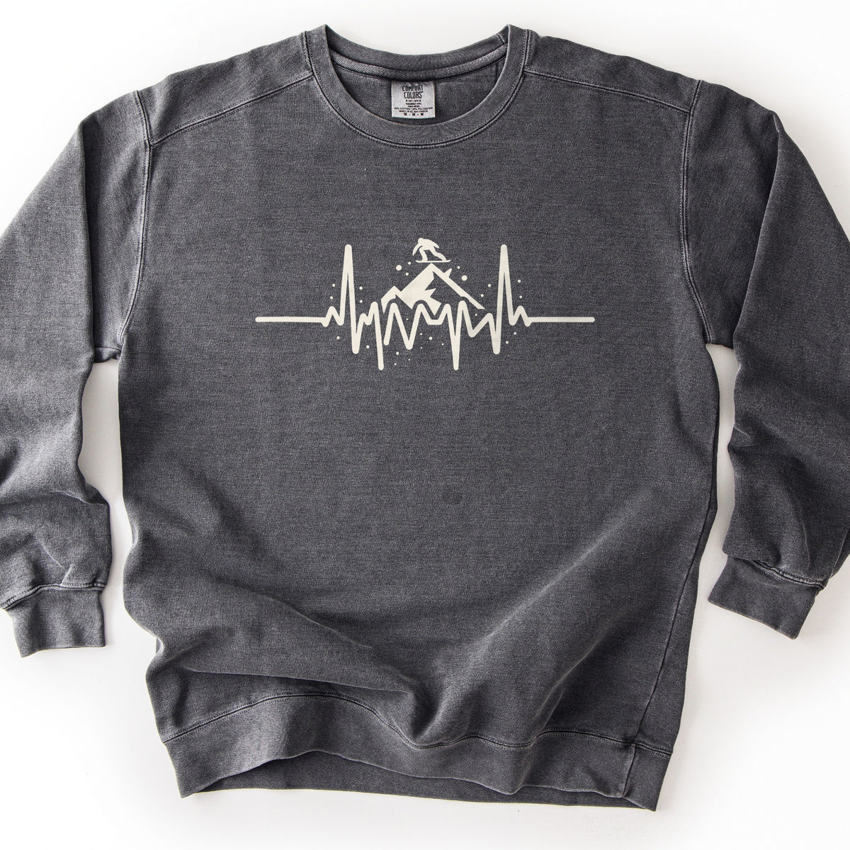 Snow Boarder Heartbeat Comfort Colors crewneck sweatshirt in Pepper, laid flat. A cream heartbeat line runs across the chest, rising into a mountain peak with a snowboarder riding on top and small dots of snow around it. No words. The sweatshirt lies on a white surface.