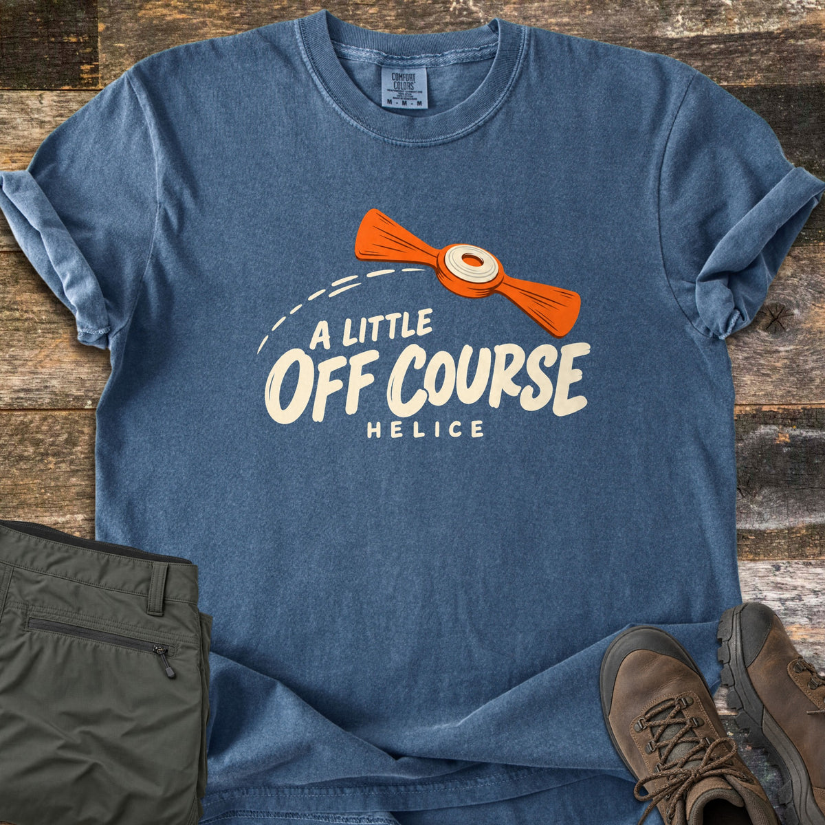 A Little Off Course Comfort Colors T-shirt in Denim, laid flat. An orange clay target flies along a dashed arc above the words A Little Off Course and Helice in cream. Sleeves are rolled on a wood plank surface beside green cargo pants and brown hiking boots.