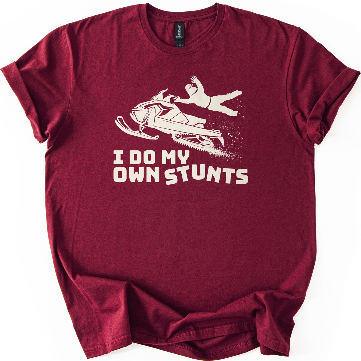 Snow Mobile Stunts Comfort Colors T-shirt in Maroon, laid flat. A cream silhouette of a snowmobile flying through the air, its rider thrown off behind it in a spray of snow, above the words I Do My Own Stunts. The shirt lies on a white background with rolled sleeves.