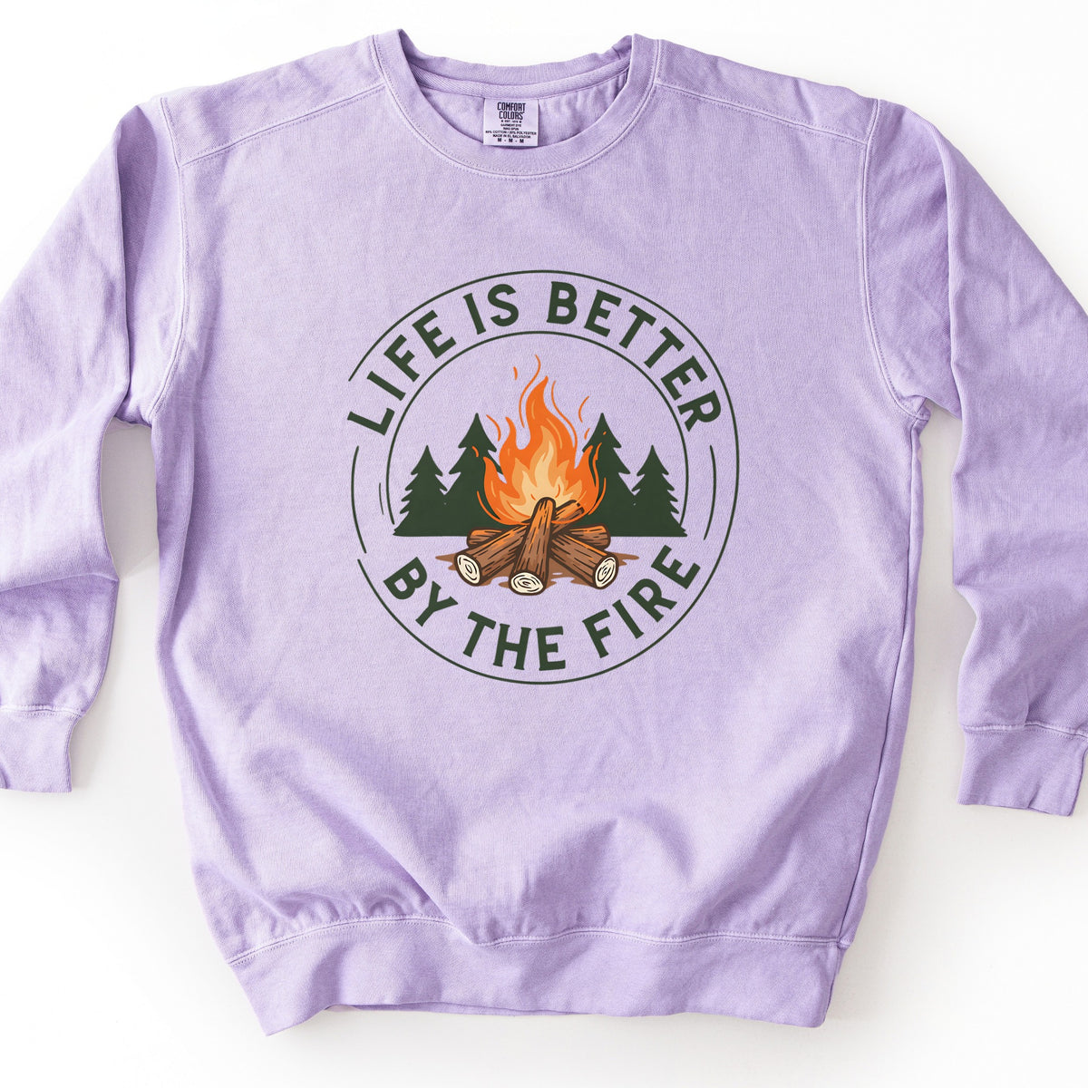 Life Is Better By The Fire Ring Comfort Colors crewneck sweatshirt in Orchid, laid flat. A circular badge shows a campfire of crossed logs with orange flames in front of dark green pine trees, ringed by the words Life Is Better By The Fire in dark green. It lies on a white surface.