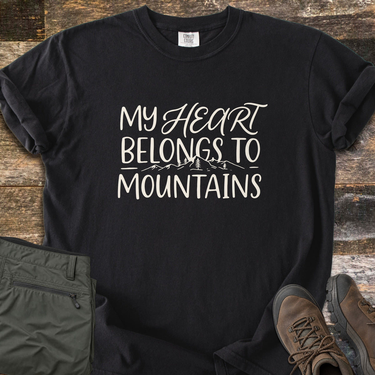 My Heart Belongs to Mountains Comfort Colors T-shirt in Black, laid flat. Cream hand lettering reads My Heart Belongs to Mountains, with Heart in script and a line drawing of mountain peaks and pine trees. Folded olive hiking pants and brown hiking boots sit on weathered wood.