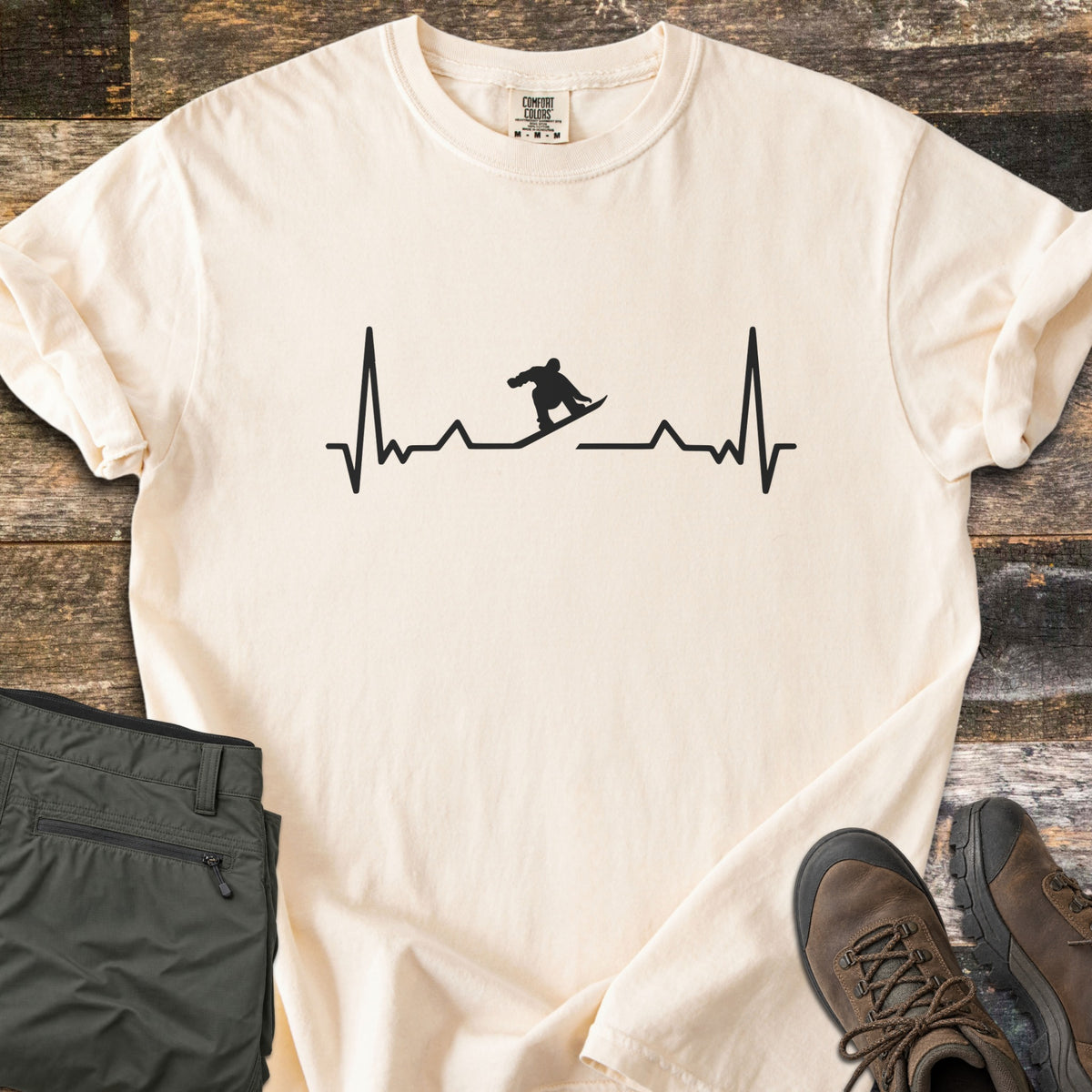 Snowboarder Heartbeat Comfort Colors T-shirt in Ivory, laid flat. A black heartbeat line runs across the chest, with a snowboarder silhouette riding the line in the middle. Sleeves are rolled, on a rustic wood floor beside folded olive pants and brown hiking boots.
