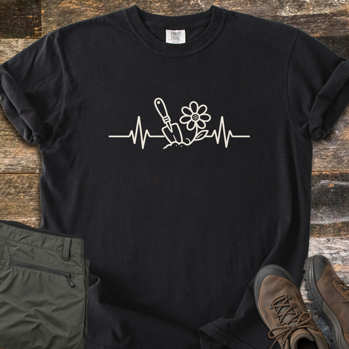 Gardening Heartbeat Comfort Colors T-shirt in Black, laid flat. A cream line drawing of a heartbeat line with a garden trowel in soil and a daisy at its center, with no words. Folded olive pants and brown hiking boots sit beside the shirt on weathered wood boards.