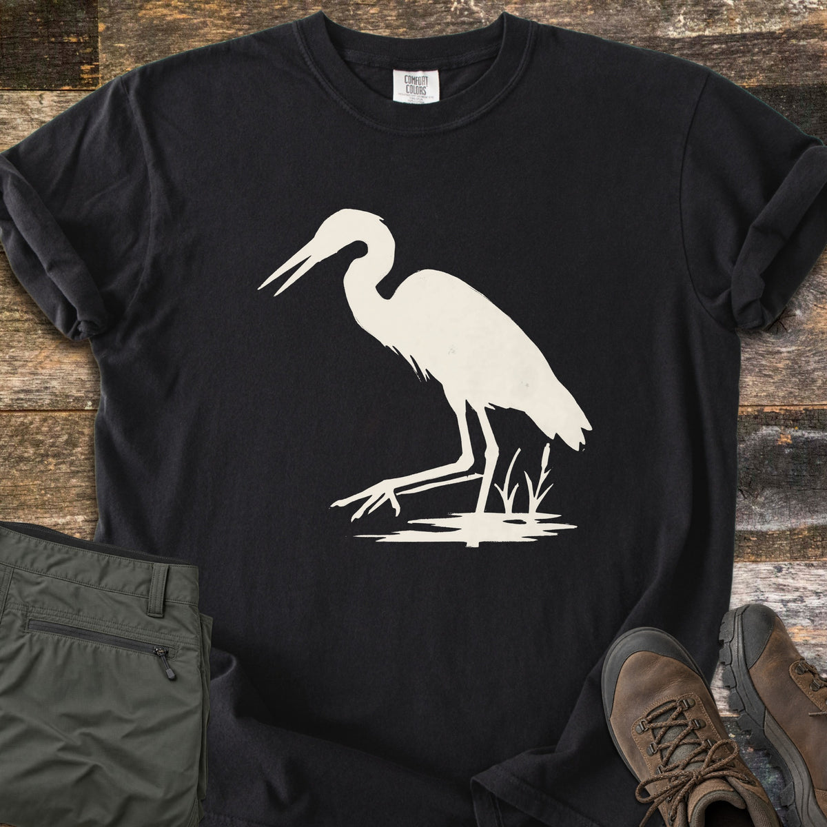 Great Blue Heron Comfort Colors T-shirt in Black, laid flat. A cream silhouette of a heron wading with its beak open, one foot raised, beside cattails over a patch of water. Sleeves rolled, on weathered wood with olive hiking pants and brown hiking boots.