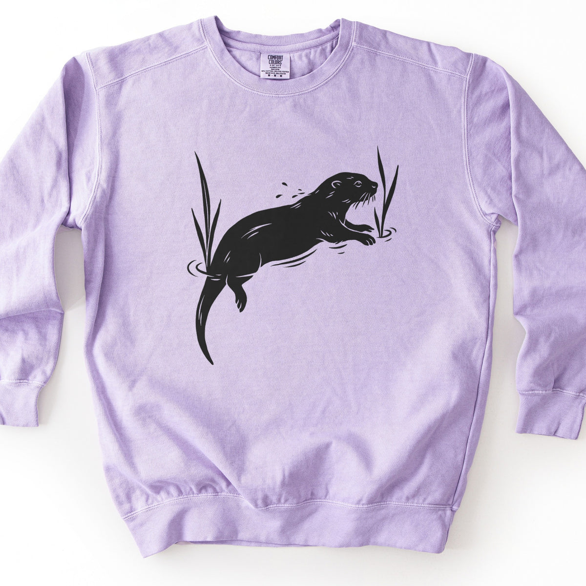 Swimming Otter Comfort Colors crewneck sweatshirt in Orchid, laid flat. A black block-print style otter swims with its head raised, splashing droplets, between two tall reeds with ripples around them. No words are printed. The sweatshirt lies on a white background.