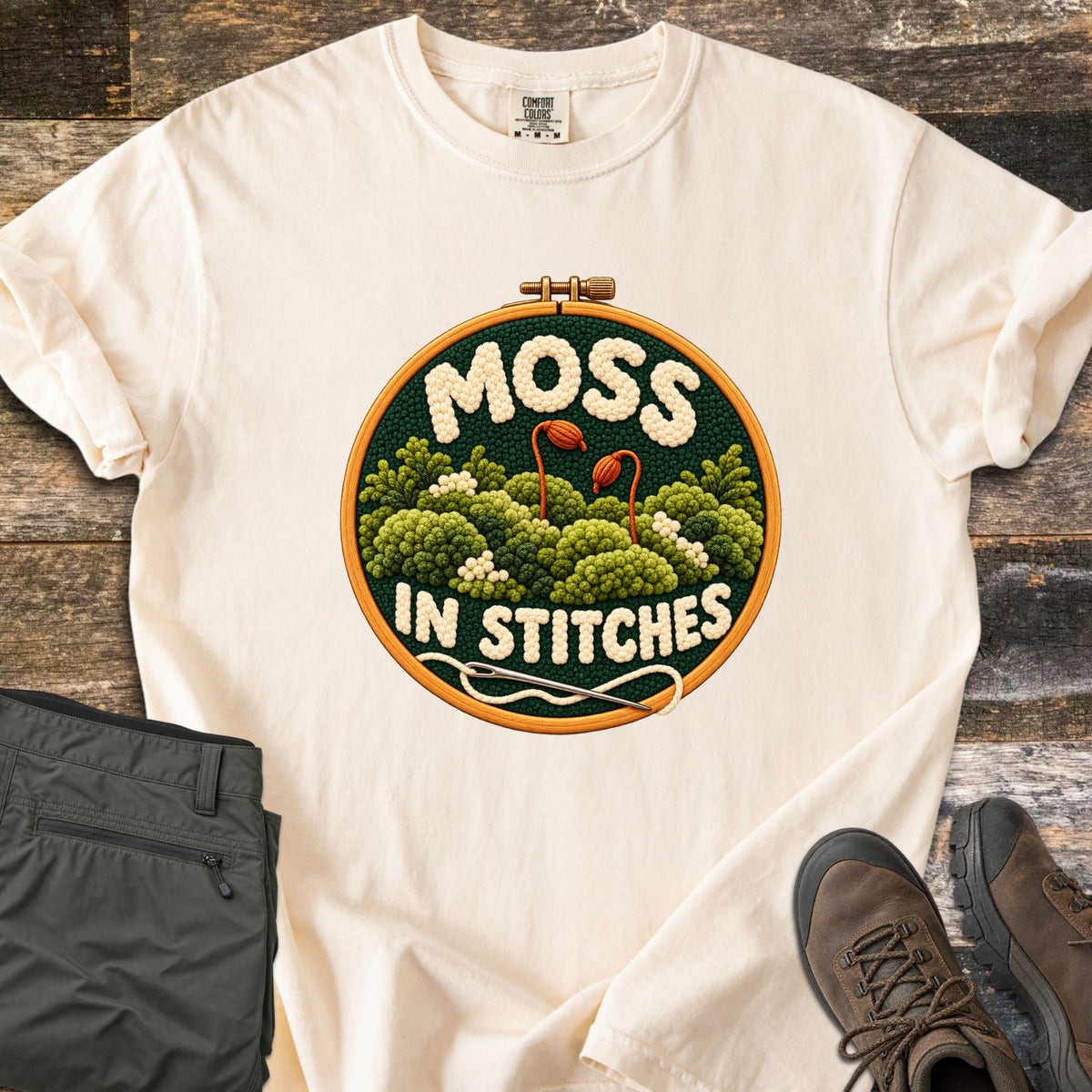 Moss in Stitches Comfort Colors T-shirt in Ivory, laid flat. An embroidery hoop holds stitched green moss with two orange spore capsules, the word Moss above and In Stitches below, plus a needle and thread. Folded olive pants and brown hiking boots lie beside it on weathered wood.
