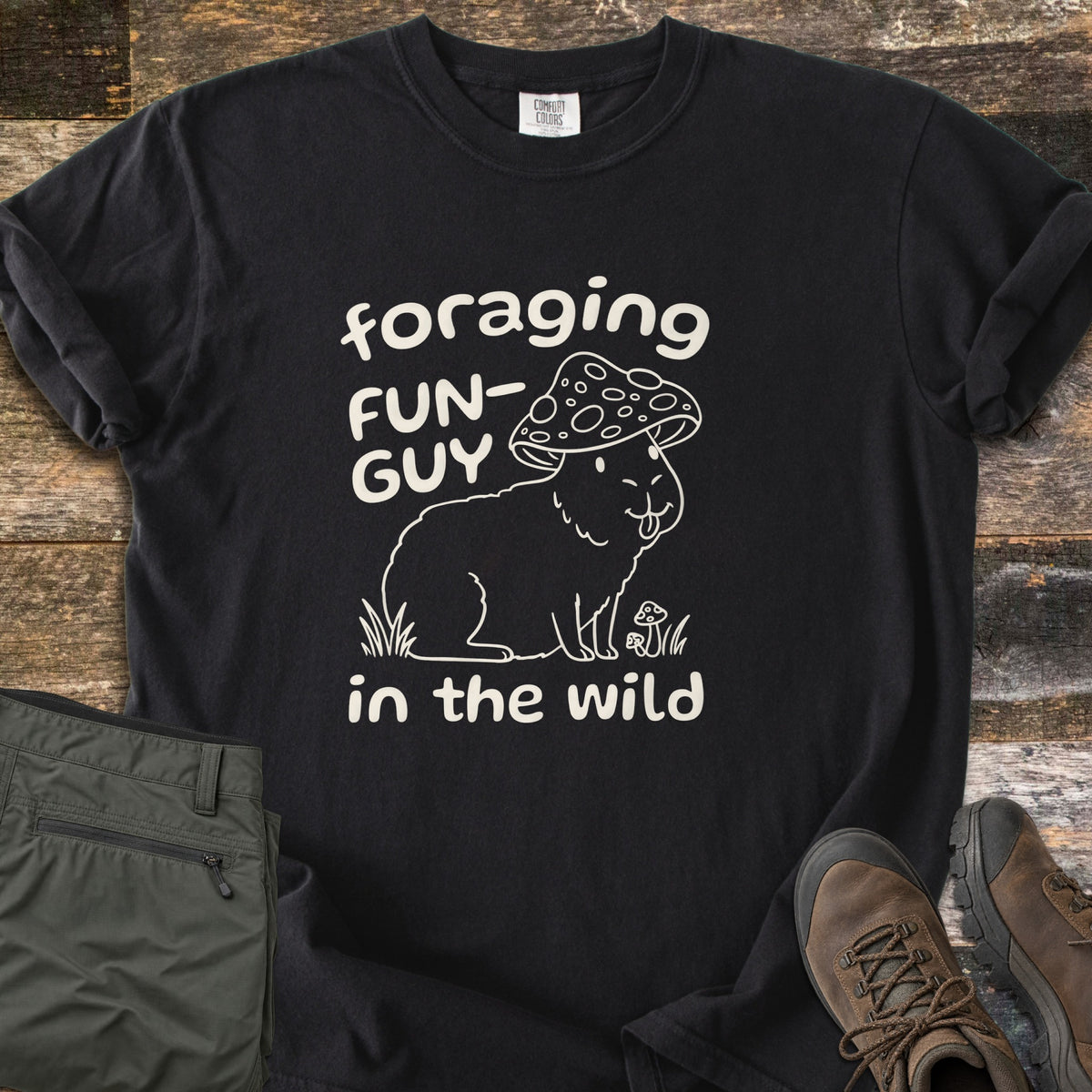 Foraging Fun-Guy in the Wild Comfort Colors T-shirt in Black, laid flat. A cream line drawing of a capybara wearing a spotted mushroom cap, tongue out, sits in grass by tiny mushrooms between the words foraging FUN-GUY in the wild. Olive hiking pants and brown boots lie on weathered wood.