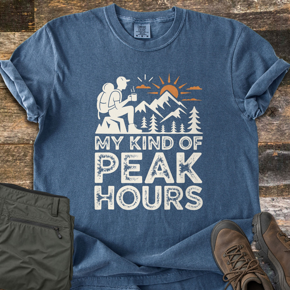 My Kind Of Peak Hours Comfort Colors T-shirt in Denim, laid flat. A white hiker with a backpack sits on a rock holding a steaming mug, facing mountains, pine trees and an orange sun, above the words My Kind Of Peak Hours. Olive hiking pants and brown boots lie on weathered wood.