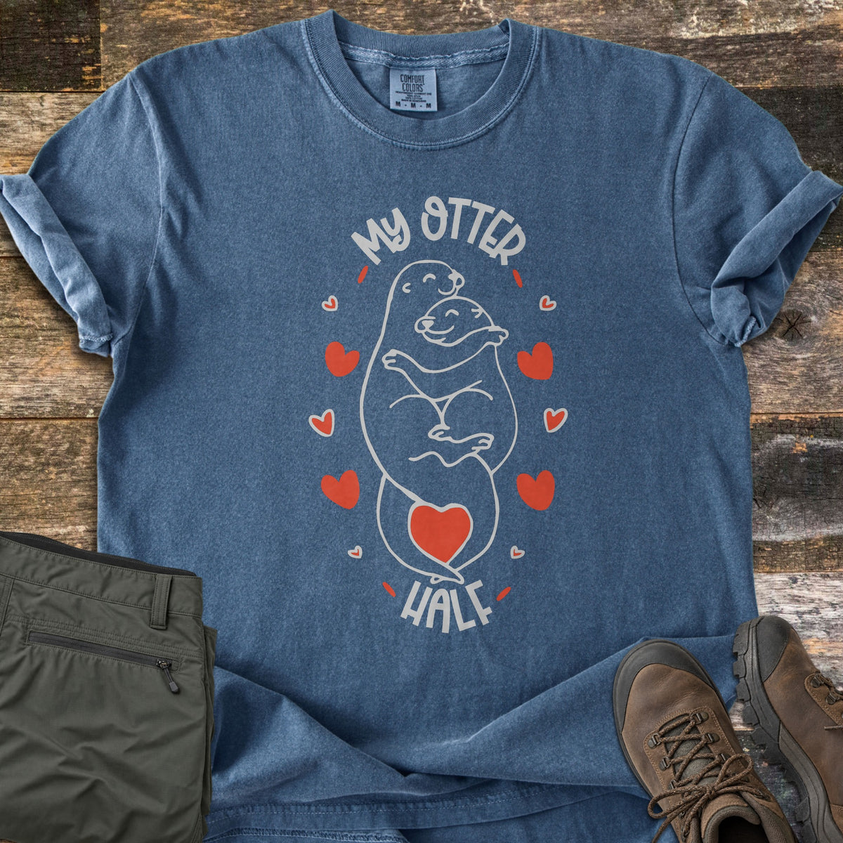My Otter Half Comfort Colors T-shirt in Denim, laid flat. A white line drawing of two otters hugging, with a red heart on the tail and red hearts around them, between the words My Otter Half. Sleeves are rolled on a wood floor beside olive pants and brown hiking boots.
