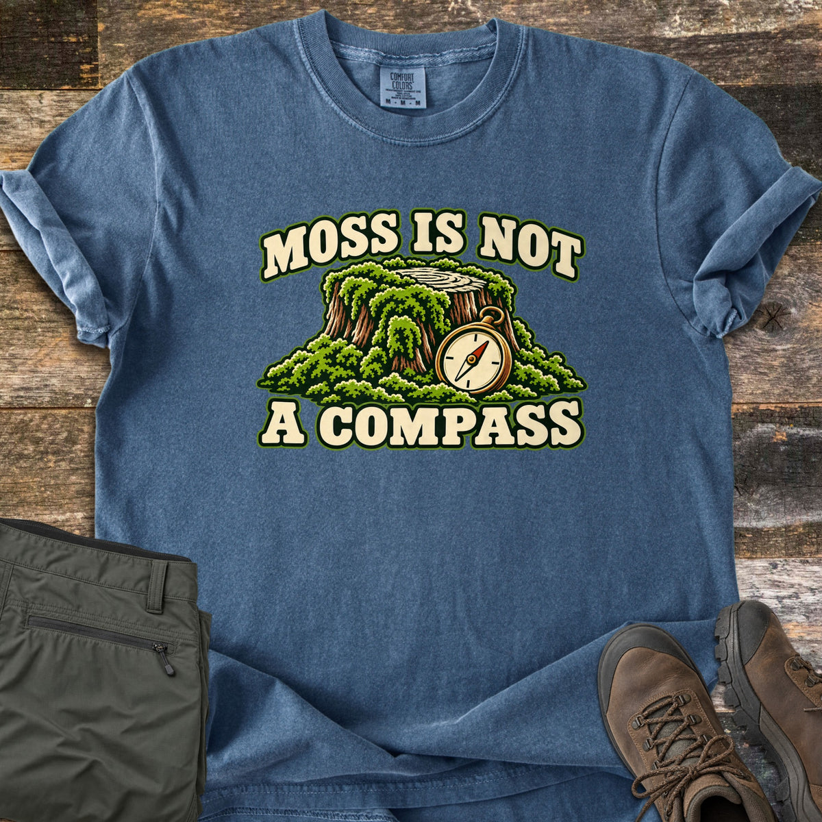 Moss Is Not a Compass Comfort Colors T-shirt in Denim, laid flat. A moss-covered tree stump with a brass compass sits between the words Moss Is Not A Compass. The shirt lies on weathered wood planks with olive hiking pants and brown hiking boots.