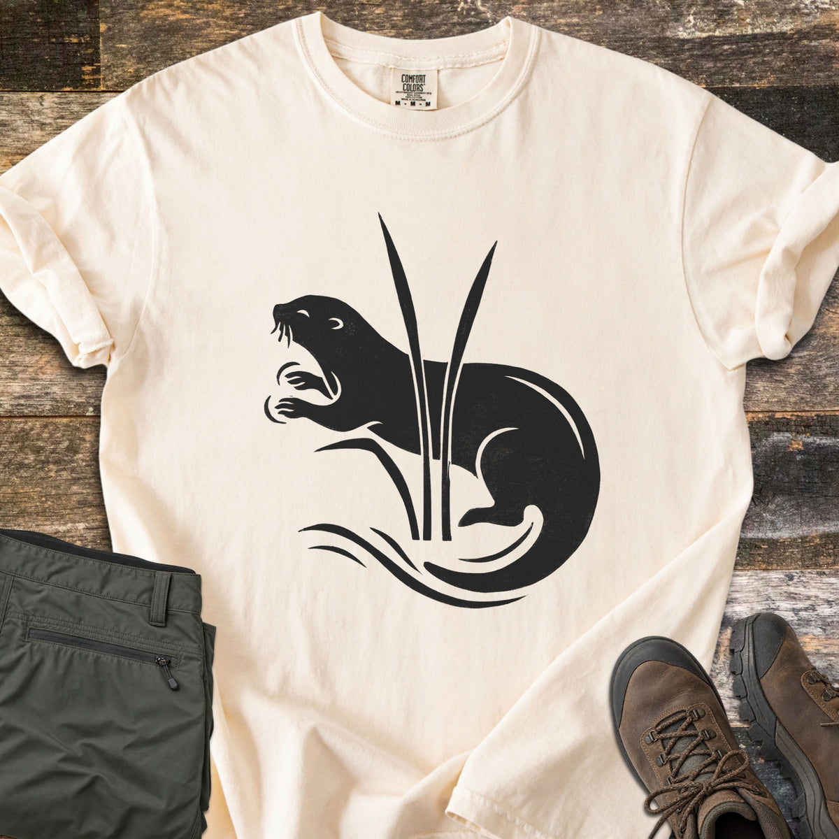 Otter In The Reeds Comfort Colors T-shirt in Ivory, laid flat. A black silhouette of an otter with open mouth and raised paws curls behind two tall reeds above wavy water lines, no words. Folded olive hiking pants and brown hiking boots lie on weathered wood.