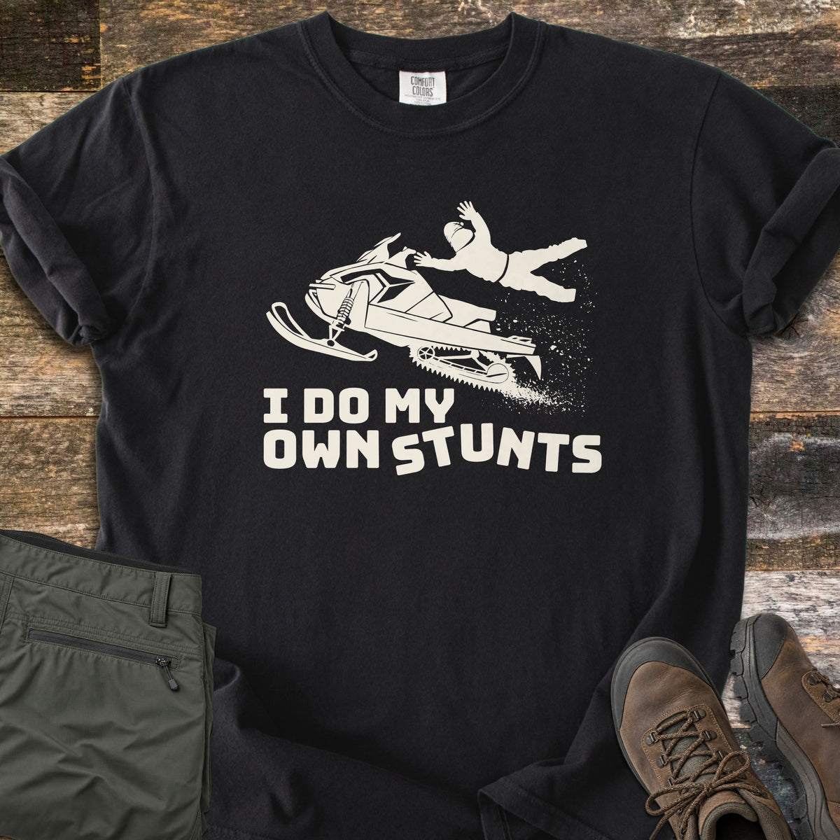 I Do My Own Stunts Comfort Colors T-shirt in Black, laid flat. A cream silhouette of a rider thrown from a snowmobile mid-jump, with snow spraying from the track, above the words I Do My Own Stunts. Olive cargo pants and brown hiking boots lie on rustic wood planks.