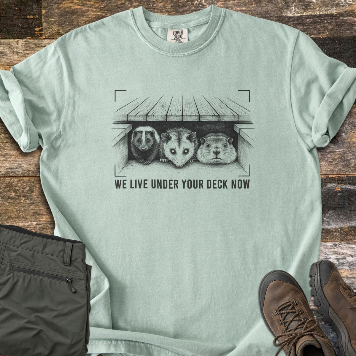We Live Under Your Deck Now Comfort Colors T-shirt in Bay, laid flat. A sketched skunk, opossum and groundhog peer from under a wooden deck inside viewfinder corners, above the words We Live Under Your Deck Now in dark gray ink. The sleeves are rolled; olive pants and brown boots sit beside it.