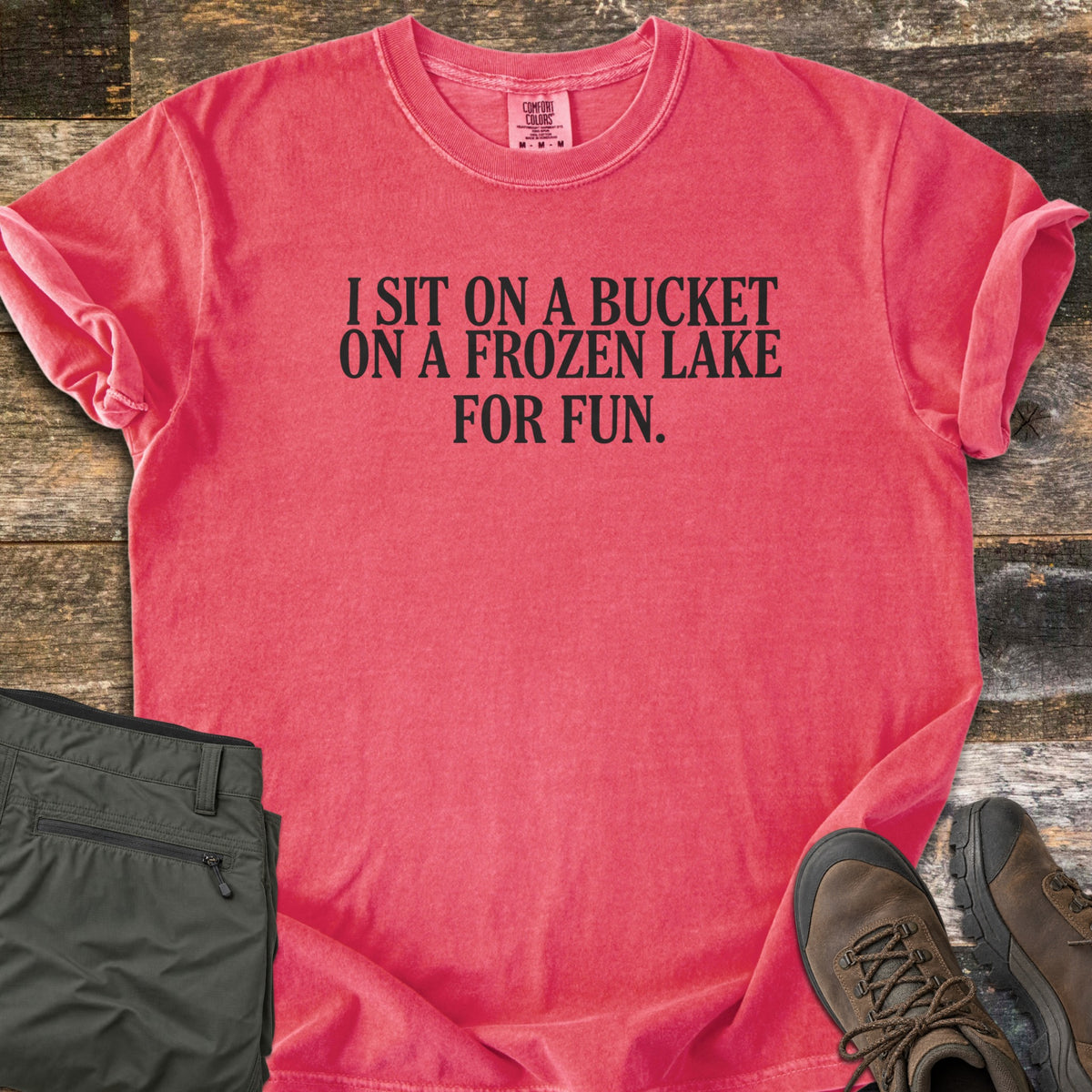 Bucket On A Frozen Lake Comfort Colors T-shirt in Watermelon, laid flat. Black serif text reads I SIT ON A BUCKET ON A FROZEN LAKE FOR FUN. The shirt has rolled sleeves and lies on weathered wood planks beside folded olive hiking pants and brown hiking boots.