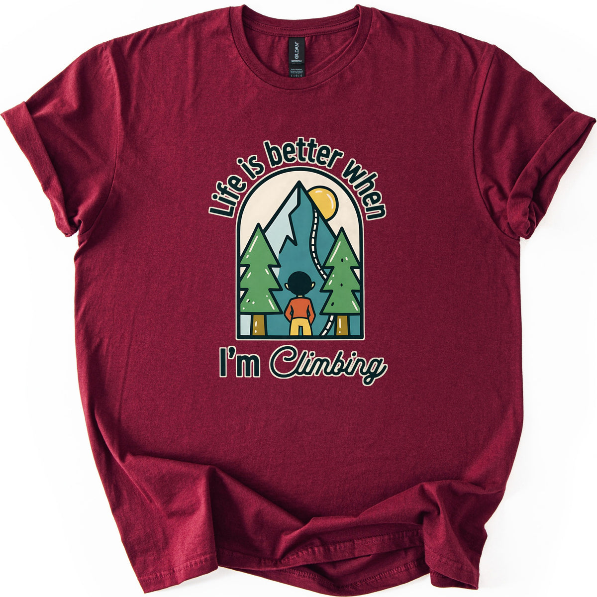 Better When I'm Climbing Comfort Colors T-shirt in Maroon, laid flat. The words Life is better when arch over a window scene of a person facing a teal mountain with a winding trail, a yellow sun and two pine trees, with I'm Climbing below. Sleeves rolled, on a white surface.