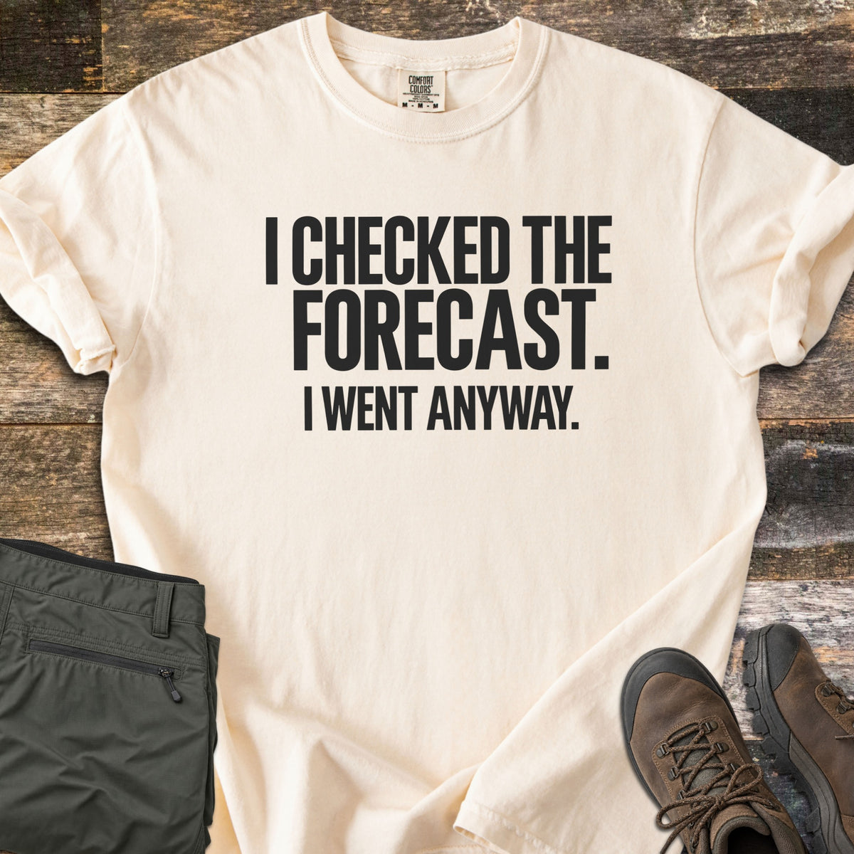 Checked The Forecast Comfort Colors T-shirt in Ivory, laid flat. Black block letters read I CHECKED THE FORECAST. I WENT ANYWAY. The sleeves are cuffed, with folded olive hiking pants and brown hiking boots beside it on weathered wood planks.