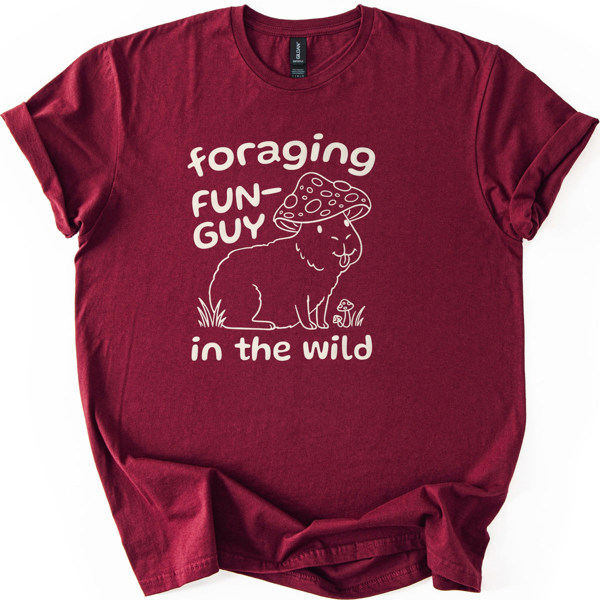 Foraging Fun-Guy Capybara Comfort Colors T-shirt in Maroon, laid flat. A cream line drawing of a winking capybara with its tongue out, wearing a spotted mushroom cap and sitting in grass by tiny mushrooms, with the words foraging FUN-GUY in the wild. Sleeves rolled, on white.