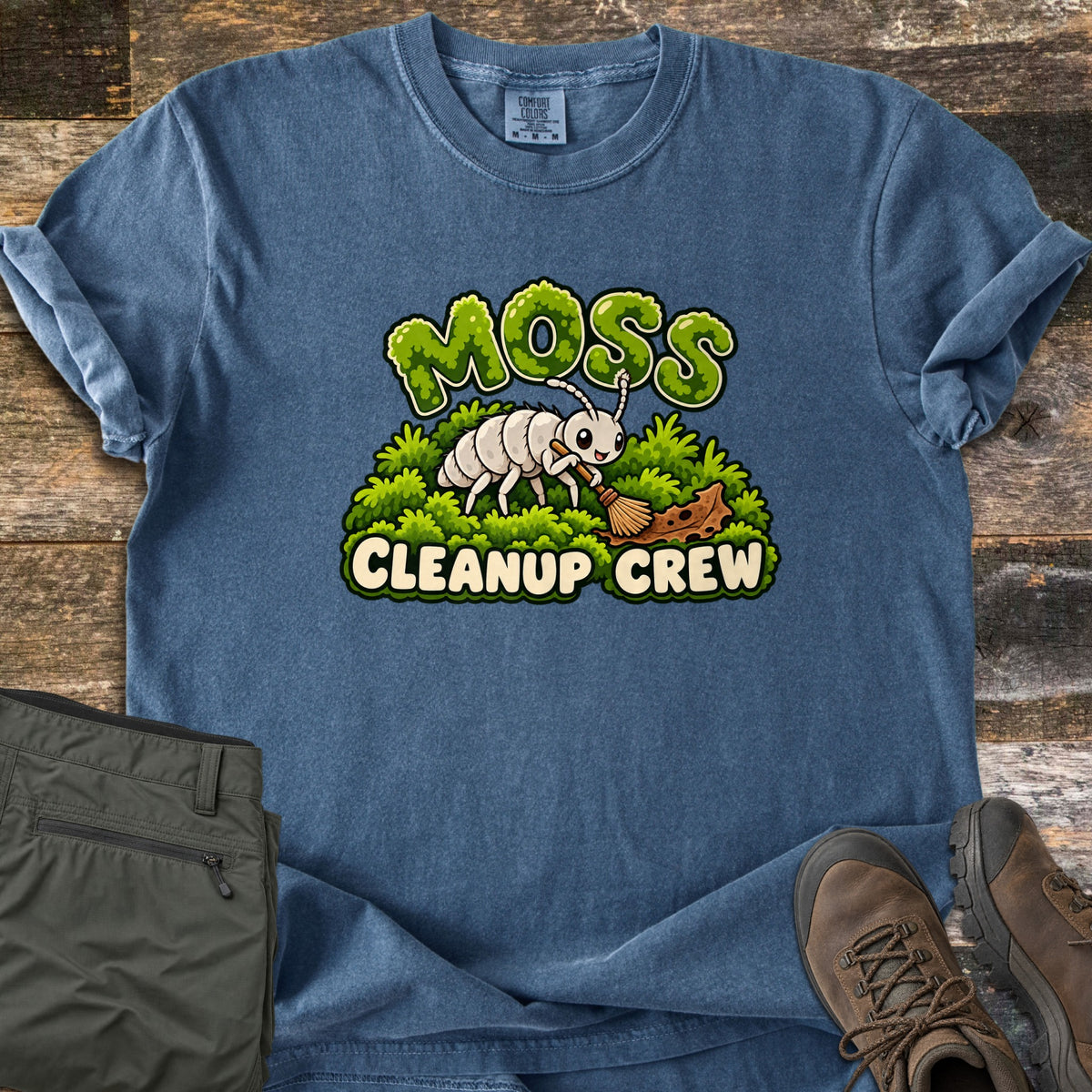 Moss Cleanup Crew Comfort Colors T-shirt in Denim, laid flat. A cartoon white bug sweeps dirt from green moss with a small broom, under the word Moss in mossy letters and above Cleanup Crew. Olive pants and brown hiking boots lie beside it on weathered wood.