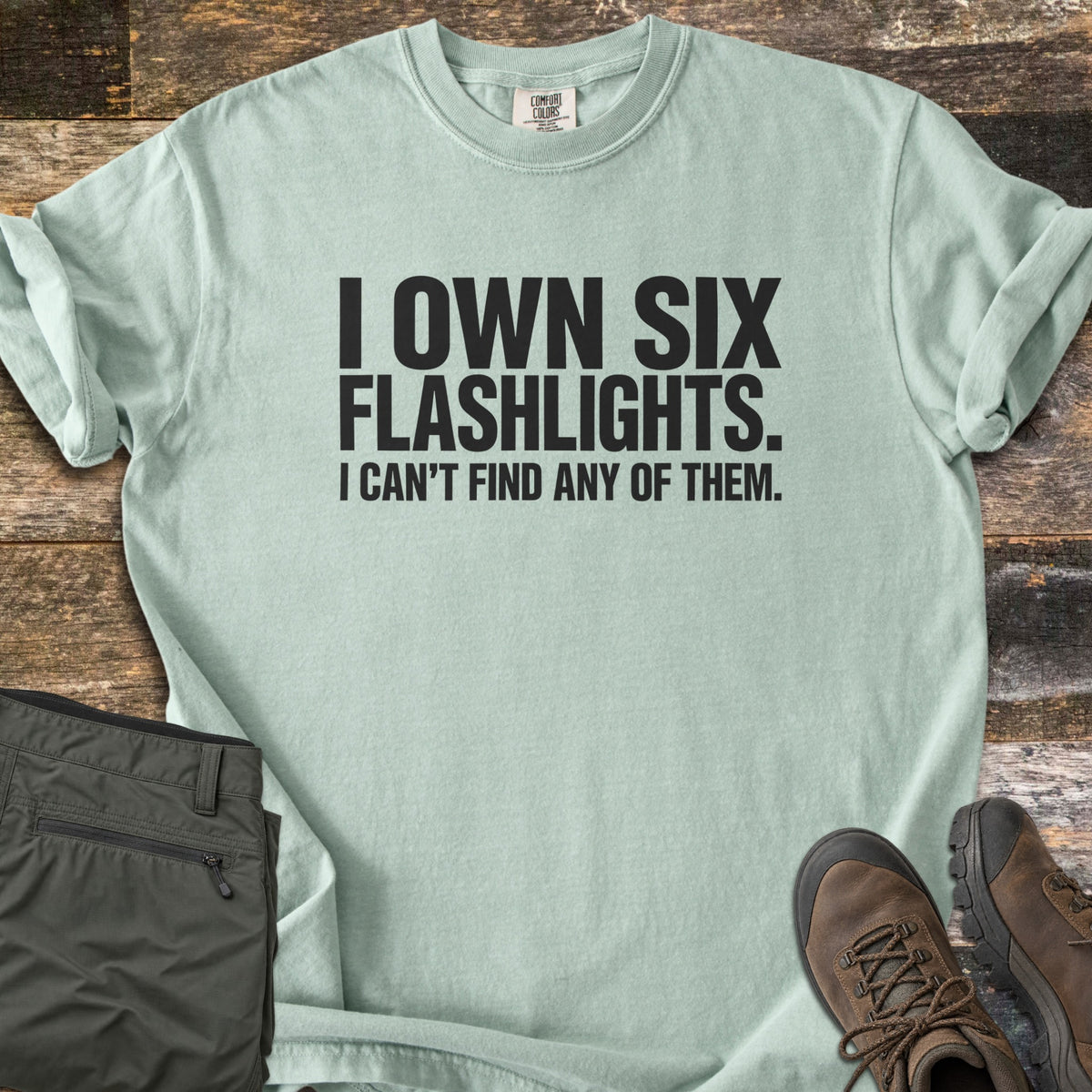 Six Flashlights Comfort Colors T-shirt in Bay, laid flat. Bold black text reads I OWN SIX FLASHLIGHTS. I CAN'T FIND ANY OF THEM. The shirt has rolled sleeves and lies on weathered wood beside folded olive cargo pants and brown hiking boots.