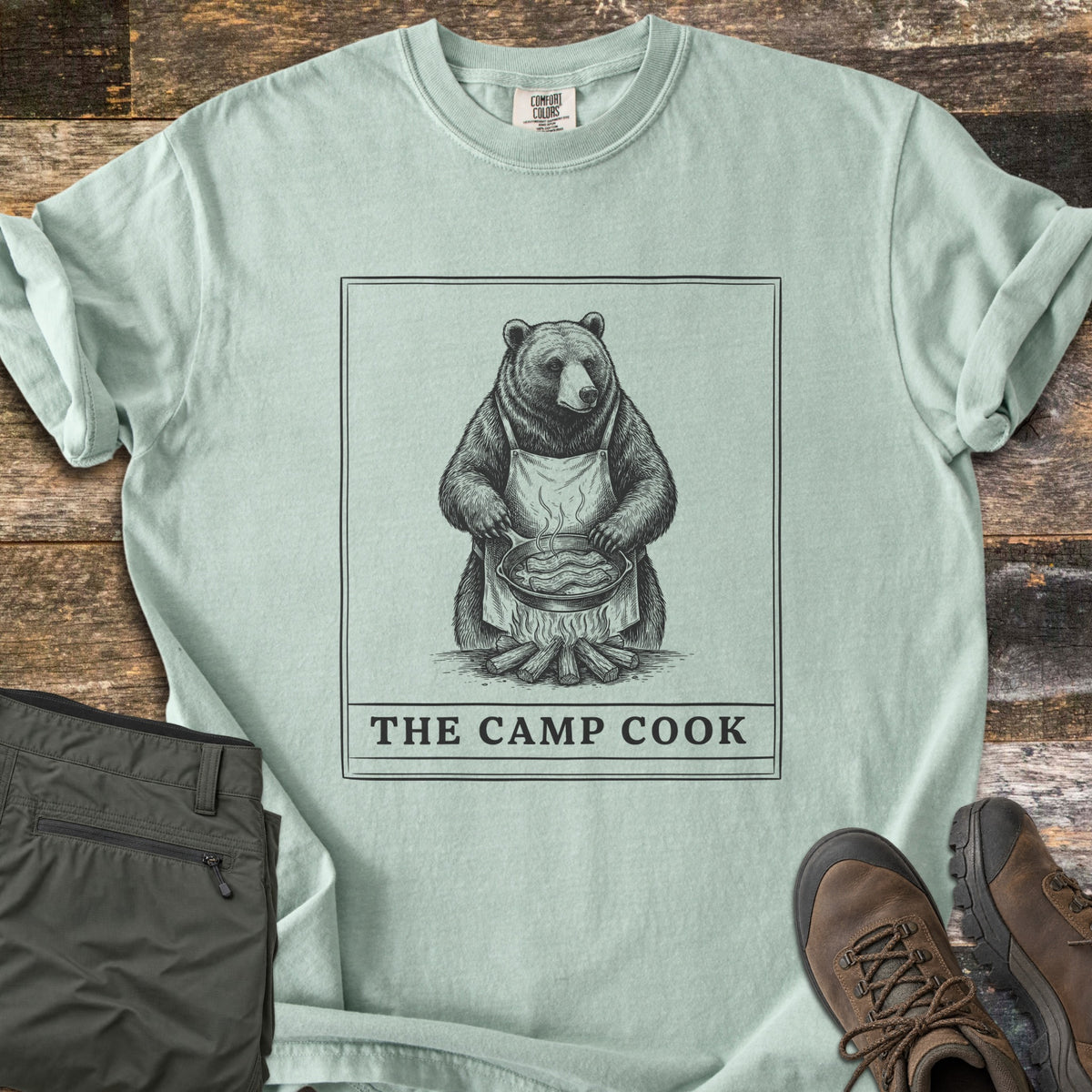 The Camp Cook Comfort Colors T-shirt in Bay, laid flat. A framed sketch of a bear in an apron frying bacon in a skillet over a campfire, above the words THE CAMP COOK. Sleeves are cuffed on weathered wood beside olive hiking pants and brown hiking boots.