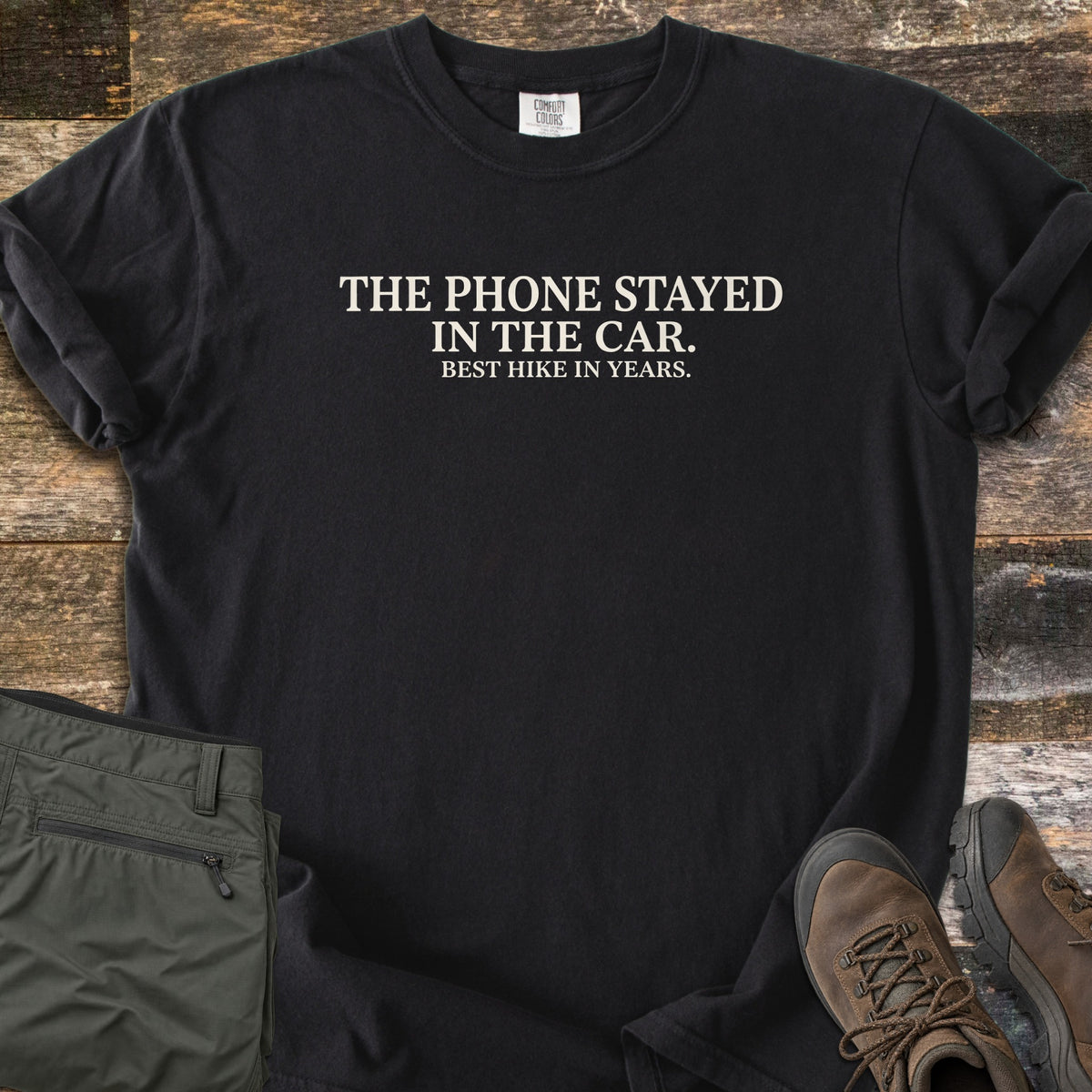Phone Stayed In The Car Comfort Colors T-shirt in Black, laid flat. Cream serif text reads THE PHONE STAYED IN THE CAR. with BEST HIKE IN YEARS. in smaller letters below. Sleeves are rolled on a wood plank surface beside olive hiking pants and brown hiking boots.