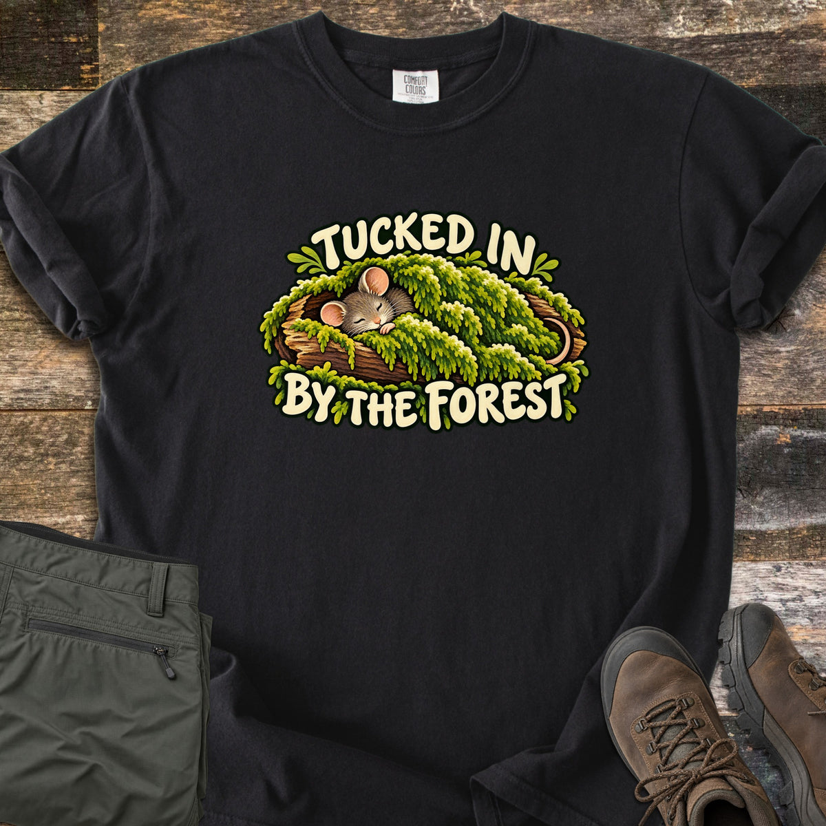 Tucked in by the Forest Comfort Colors T-shirt in Black, laid flat. A sleeping cartoon mouse lies under a blanket of green moss in a hollow log between the words Tucked In By The Forest. Folded olive hiking pants and brown hiking boots sit on weathered wood planks.