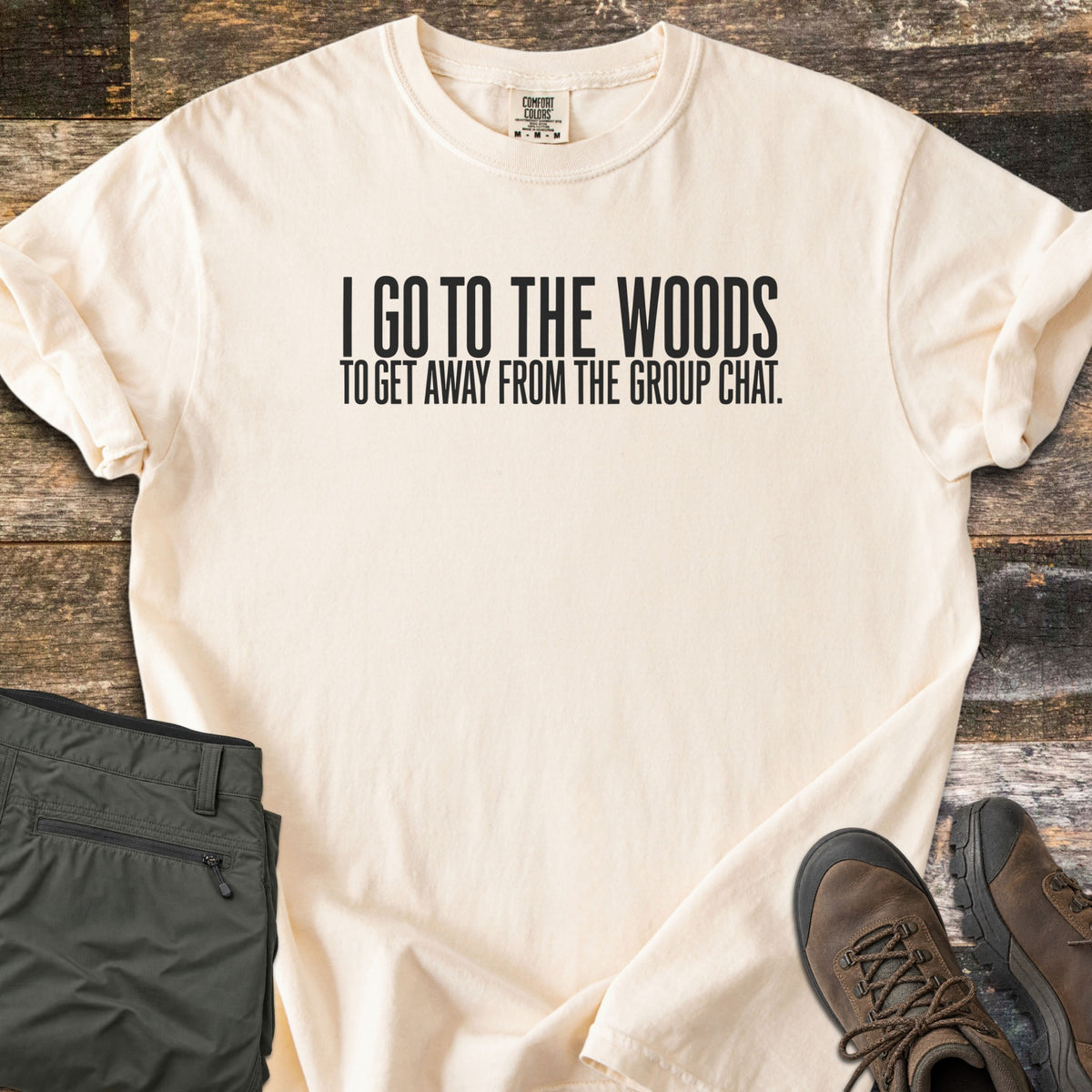 Away From The Group Chat Comfort Colors T-shirt in Ivory, laid flat. Black block letters read I GO TO THE WOODS above TO GET AWAY FROM THE GROUP CHAT. The shirt has rolled sleeves and lies on weathered wood boards beside folded olive green pants and brown hiking boots.