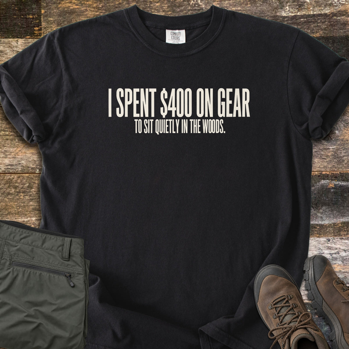 Gear To Sit Quietly Comfort Colors T-shirt in Black, laid flat. Cream text reads I SPENT $400 ON GEAR, with TO SIT QUIETLY IN THE WOODS. below. Sleeves are rolled on a rustic wood surface, beside folded olive hiking pants and brown hiking boots.