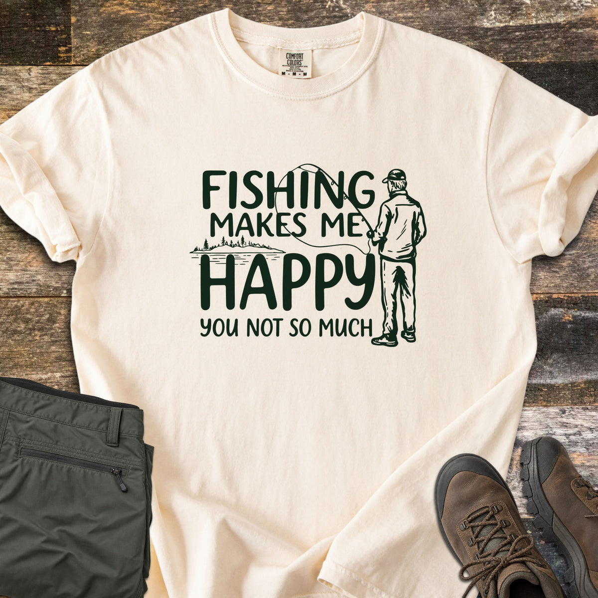 Fishing Makes Me Happy Comfort Colors T-shirt in Ivory, laid flat. Dark green print of a man in a cap casting a fishing rod by a tree-lined lake, with the words Fishing Makes Me Happy You Not So Much. Laid on weathered wood with olive pants and brown hiking boots.
