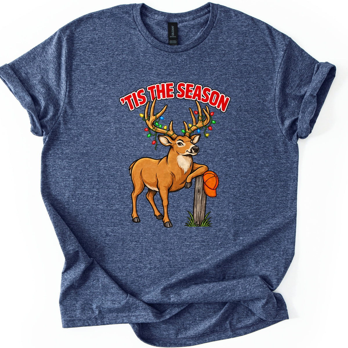 Buck Tis The Season Comfort Colors T-shirt in Heather Navy, laid flat. Red arched text reads 'TIS THE SEASON above a cartoon buck with colored Christmas lights strung on its antlers, leaning on a wooden post that holds an orange hunting cap. Sleeves rolled, on a white surface.
