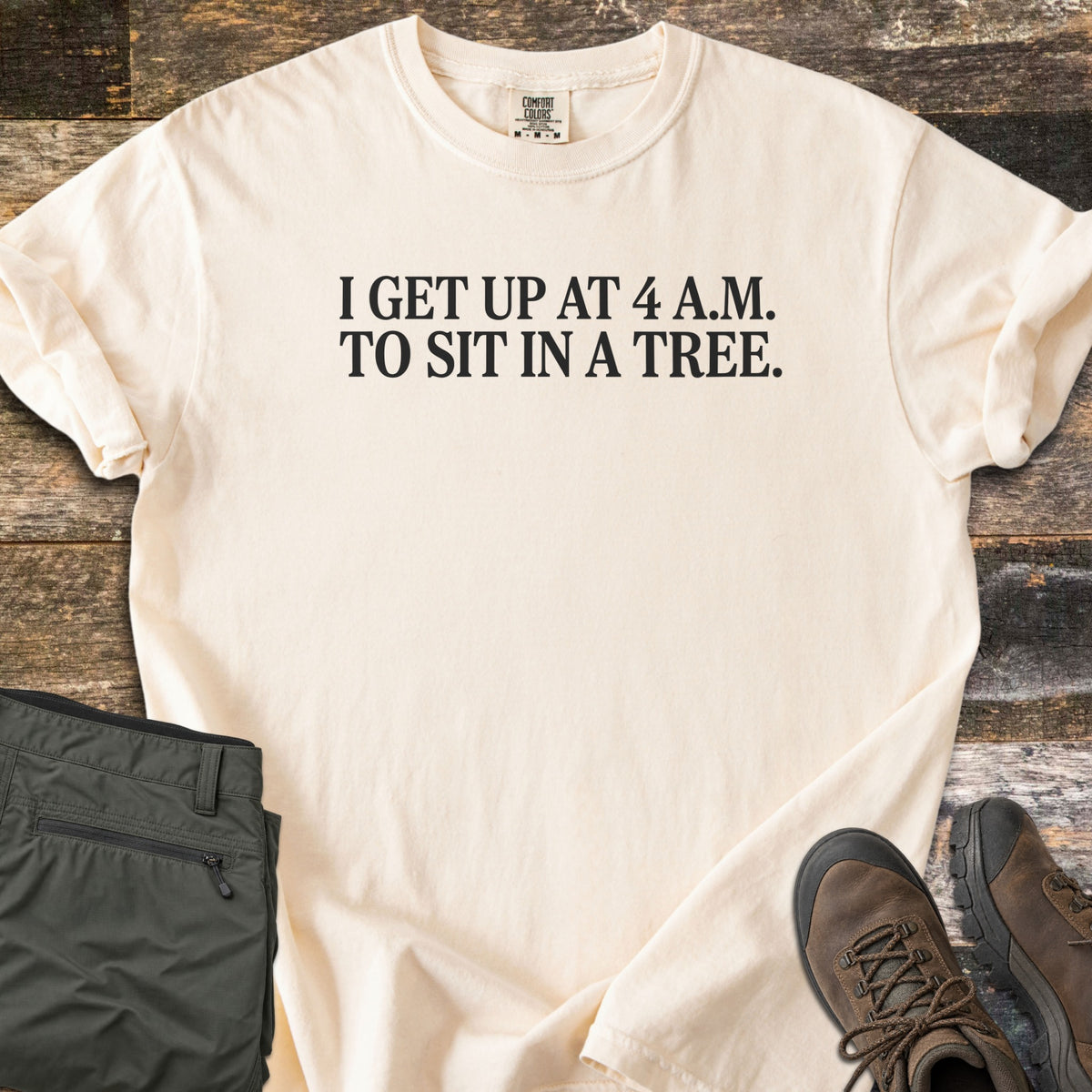 Sit In A Tree Comfort Colors T-shirt in Ivory, laid flat. Black serif text reads I GET UP AT 4 A.M. TO SIT IN A TREE. across the chest. The shirt lies on weathered wood planks beside folded olive green cargo pants and brown leather hiking boots.