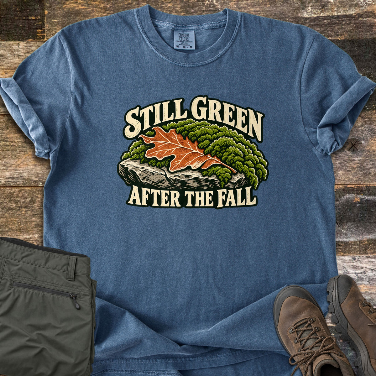 Still Green After the Fall Comfort Colors T-shirt in Denim, laid flat. An orange oak leaf rests on a mossy green rock between the words Still Green and After the Fall. Olive hiking pants and brown hiking boots sit beside it on weathered wood planks.