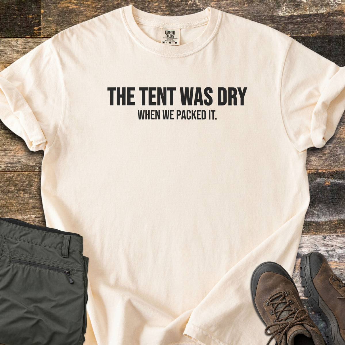 The Tent Was Dry Comfort Colors T-shirt in Ivory, laid flat. Bold black capital letters read THE TENT WAS DRY, with smaller text below reading WHEN WE PACKED IT. The shirt lies on weathered wood planks with rolled sleeves, beside folded olive hiking pants and brown hiking boots.
