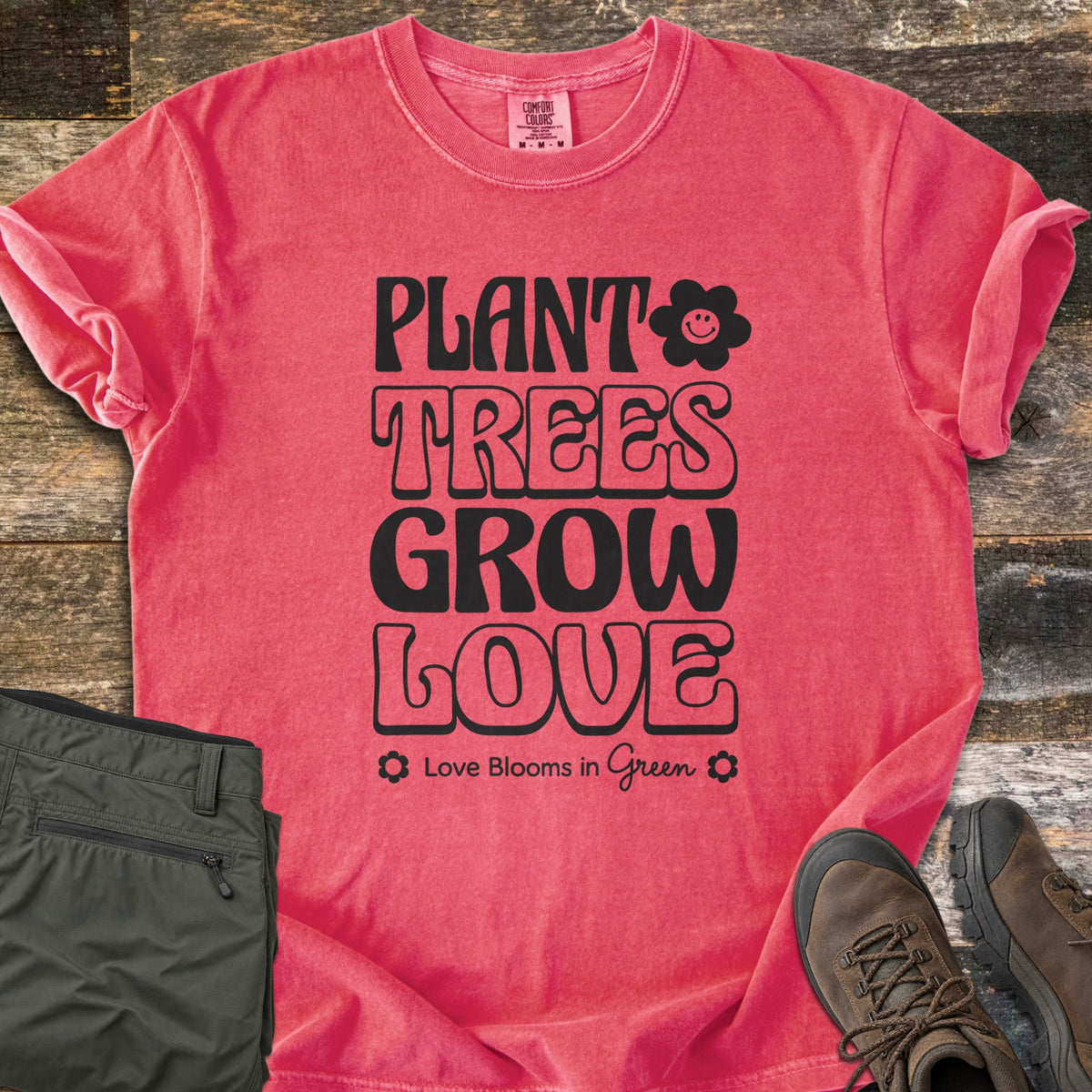 Plant Trees Grow Love Comfort Colors T-shirt in Watermelon, laid flat. Black retro bubble letters read Plant Trees Grow Love beside a smiling daisy, above Love Blooms in Green. Sleeves cuffed on weathered wood with folded olive hiking pants and brown hiking boots.