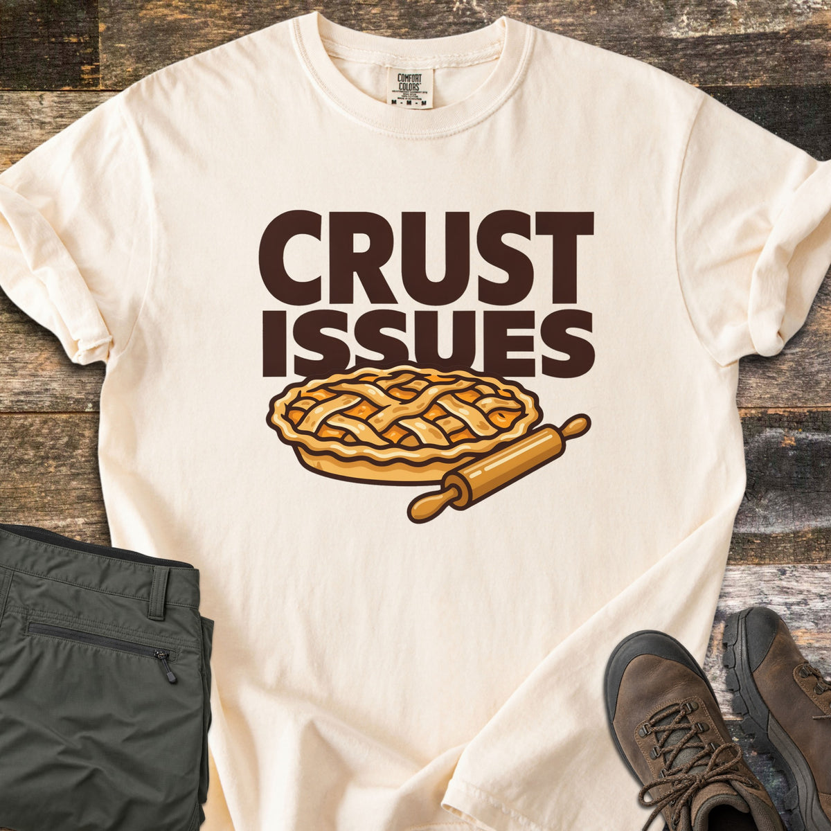 Crust Issues Comfort Colors T-shirt in Ivory, laid flat. The words CRUST ISSUES in dark brown block letters sit above a cartoon lattice-top pie and a wooden rolling pin. Olive pants and brown hiking boots lie beside it on a rustic wood surface.