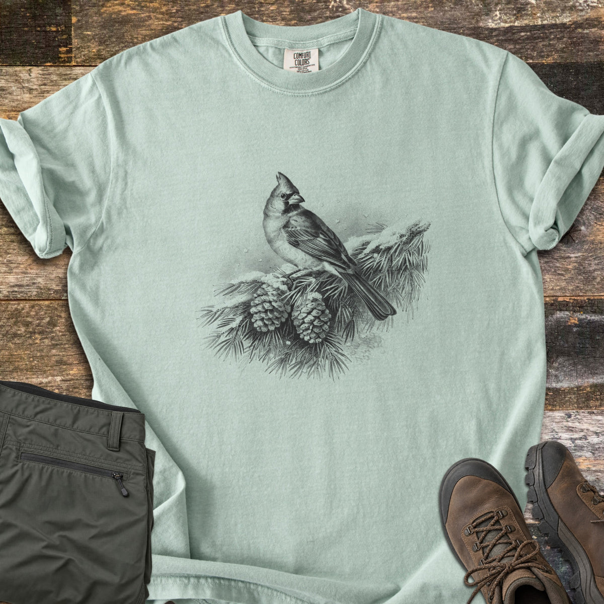 Cardinal On A Snowy Bough Comfort Colors T-shirt in Bay, laid flat. A grayscale drawing of a cardinal perched on a snow-covered pine branch with two pinecones, with no words. Sleeves rolled, on weathered wood planks beside olive hiking pants and brown hiking boots.