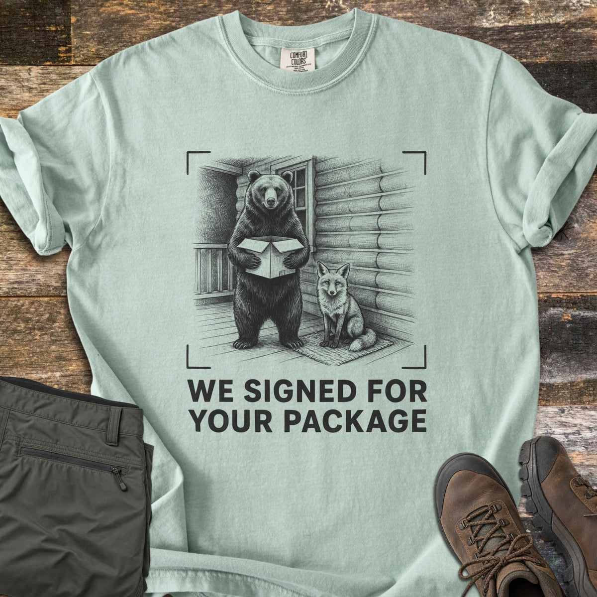 We Signed For Your Package Comfort Colors T-shirt in Bay, laid flat. A black-ink sketch shows a bear holding an open box beside a seated fox on a log cabin porch, framed by camera corner marks above the words We Signed For Your Package. Olive pants and brown hiking boots sit on wood planks.