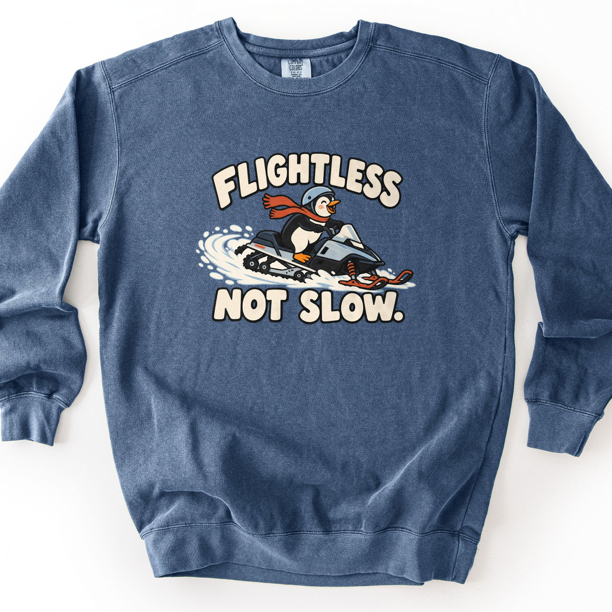 Flightless Not Slow Comfort Colors crewneck sweatshirt in Denim, laid flat. A cartoon penguin in a gray helmet and red scarf rides a snowmobile through a spray of snow between the words Flightless and Not Slow. in cream lettering. Shown on a white background.