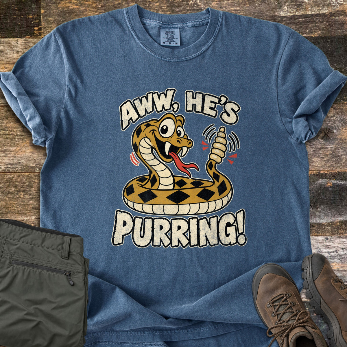 Aww He's Purring Comfort Colors T-shirt in Denim, laid flat. A cartoon rattlesnake with wide eyes and its tongue out shakes its rattle between the words Aww, He's Purring! Sleeves rolled, on weathered wood beside olive cargo pants and brown hiking boots.