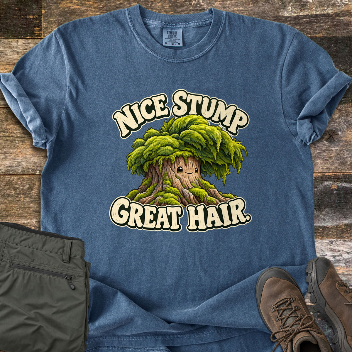 Nice Stump Great Hair Comfort Colors T-shirt in Denim, laid flat. A cartoon tree stump with a small smiling face and a bushy green mossy canopy like hair, between the words Nice Stump Great Hair. Olive hiking pants and brown hiking boots lie beside it on weathered wood planks.