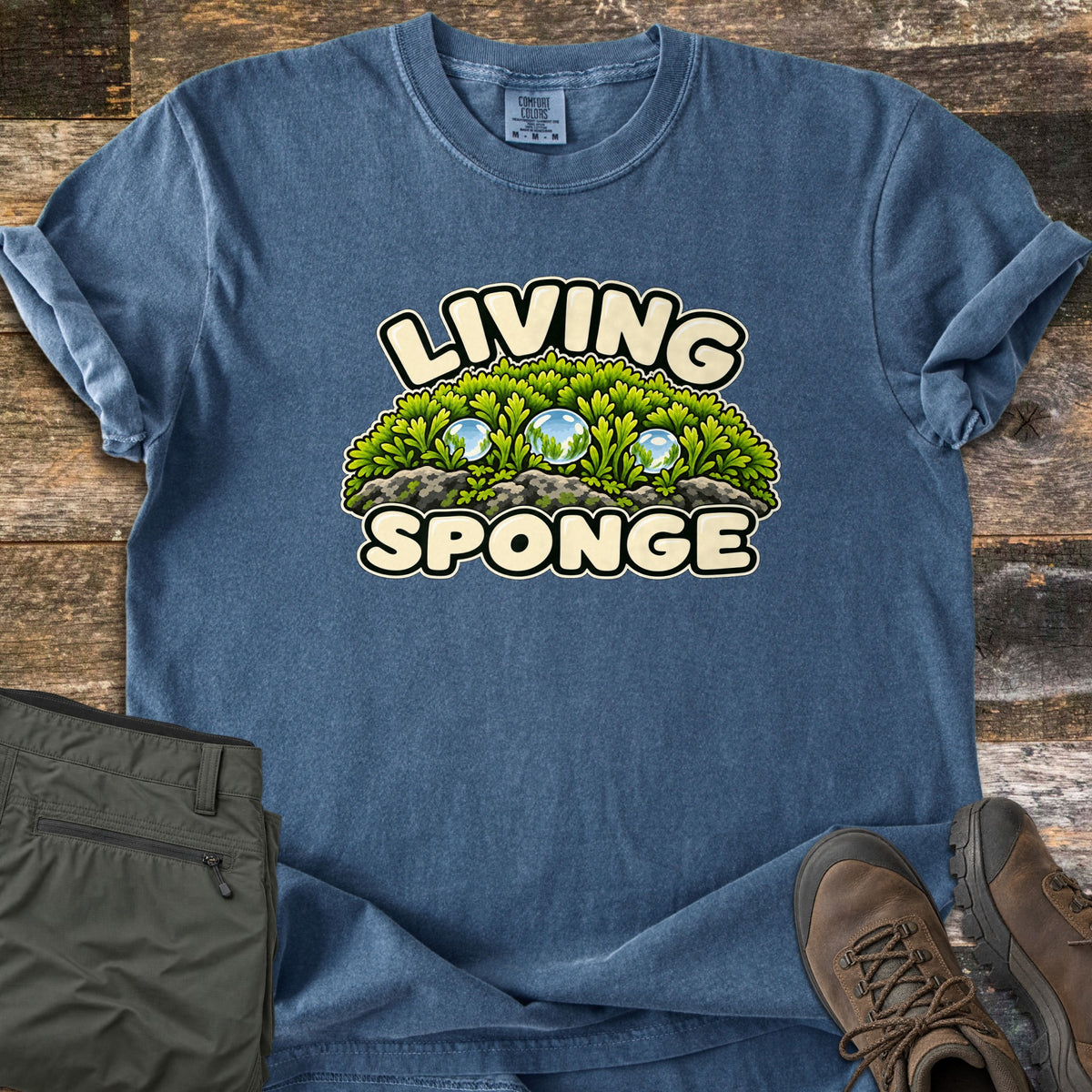 Living Sponge Comfort Colors T-shirt in Denim, laid flat. A dome of bright green moss on gray rocks holds three clear water droplets, between the words Living Sponge. Shown on weathered wood with folded olive hiking pants and brown hiking boots.