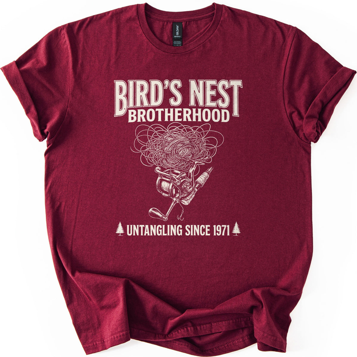 Birds Nest Brotherhood Comfort Colors T-shirt in Maroon, laid flat. Cream print reads Bird's Nest Brotherhood above a tangled mess of fishing line over a spinning reel, rod and hook, then Untangling Since 1971 between two pine trees. Sleeves cuffed, on a white backdrop.