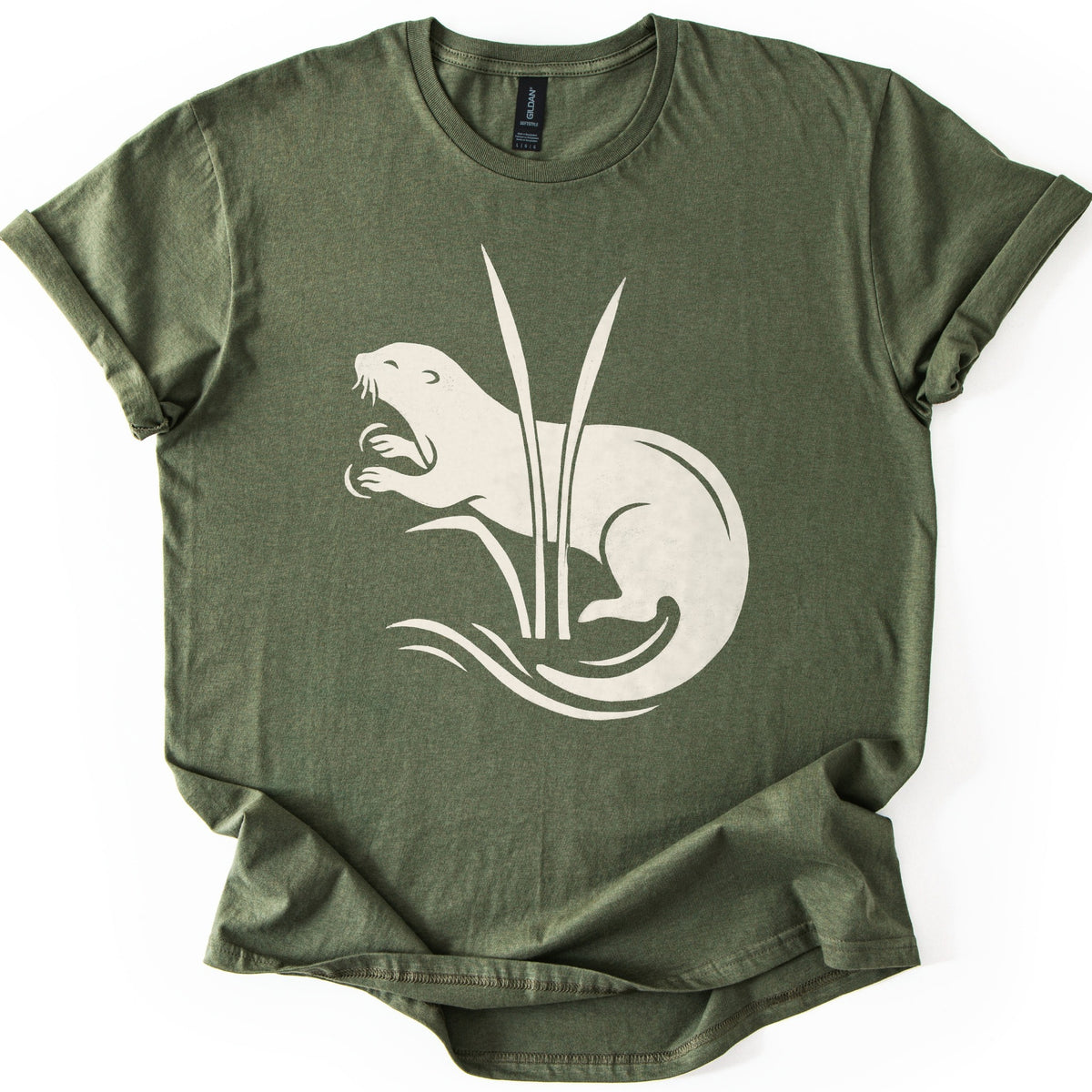Otter In The Reeds Comfort Colors T-shirt in Military Green, laid flat. A cream silhouette of an otter with open mouth, whiskers and raised paws curls behind two tall reeds above swirling water lines. No words. The shirt lies on a white surface with sleeves rolled.