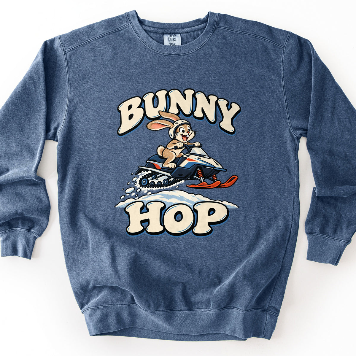 Bunny Hop Comfort Colors crewneck sweatshirt in Denim, laid flat. A cartoon rabbit in a white helmet rides a navy, white and orange snowmobile over a snowbank, between the words Bunny Hop in cream bubble letters. The sweatshirt lies on a plain white background.
